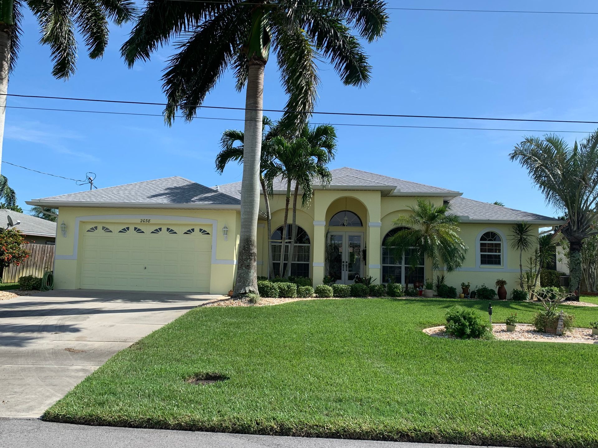 Single-story beige house with a two-car garage, palm trees, and a manicured lawn under a blue sky