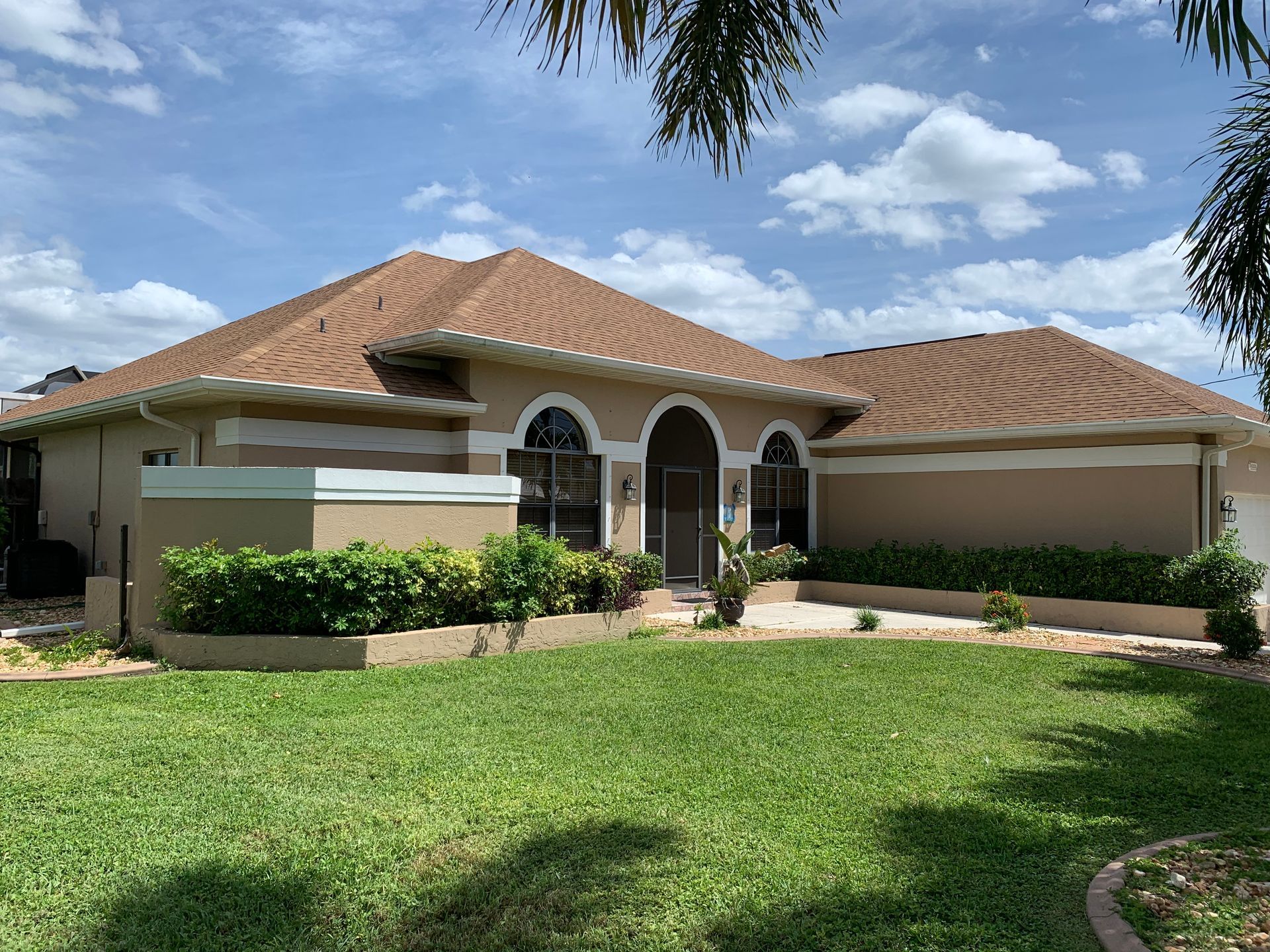 Single-story suburban house with tan stucco, red tile roof, arched entry, and a green lawn under a blue sky.