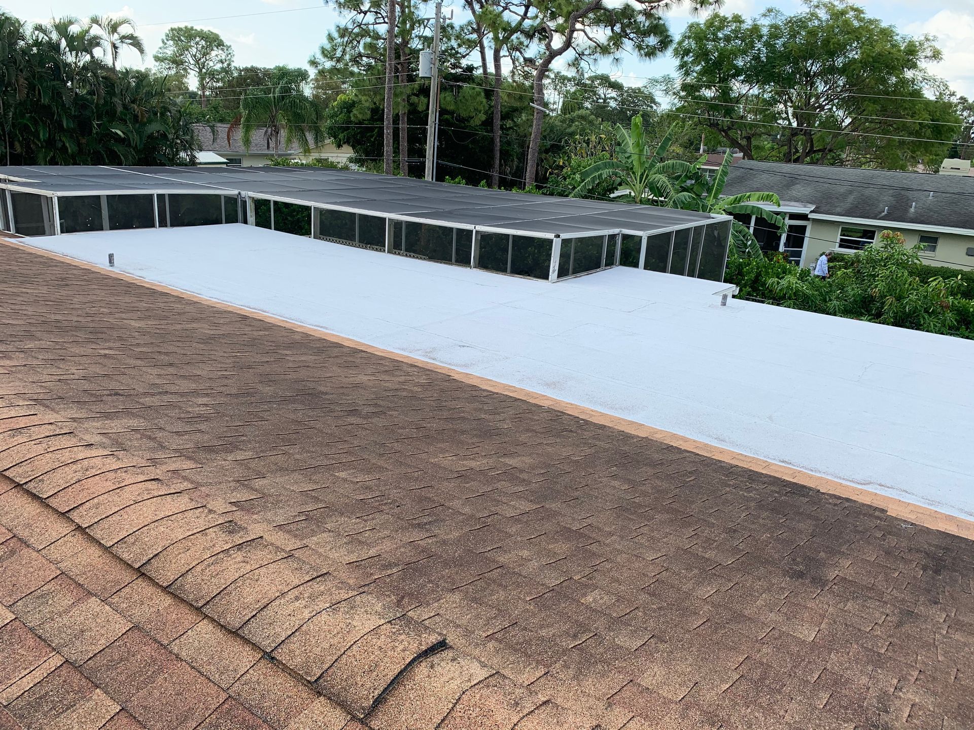 Rooftop with weathered shingles beside a large white roof and trees in the background