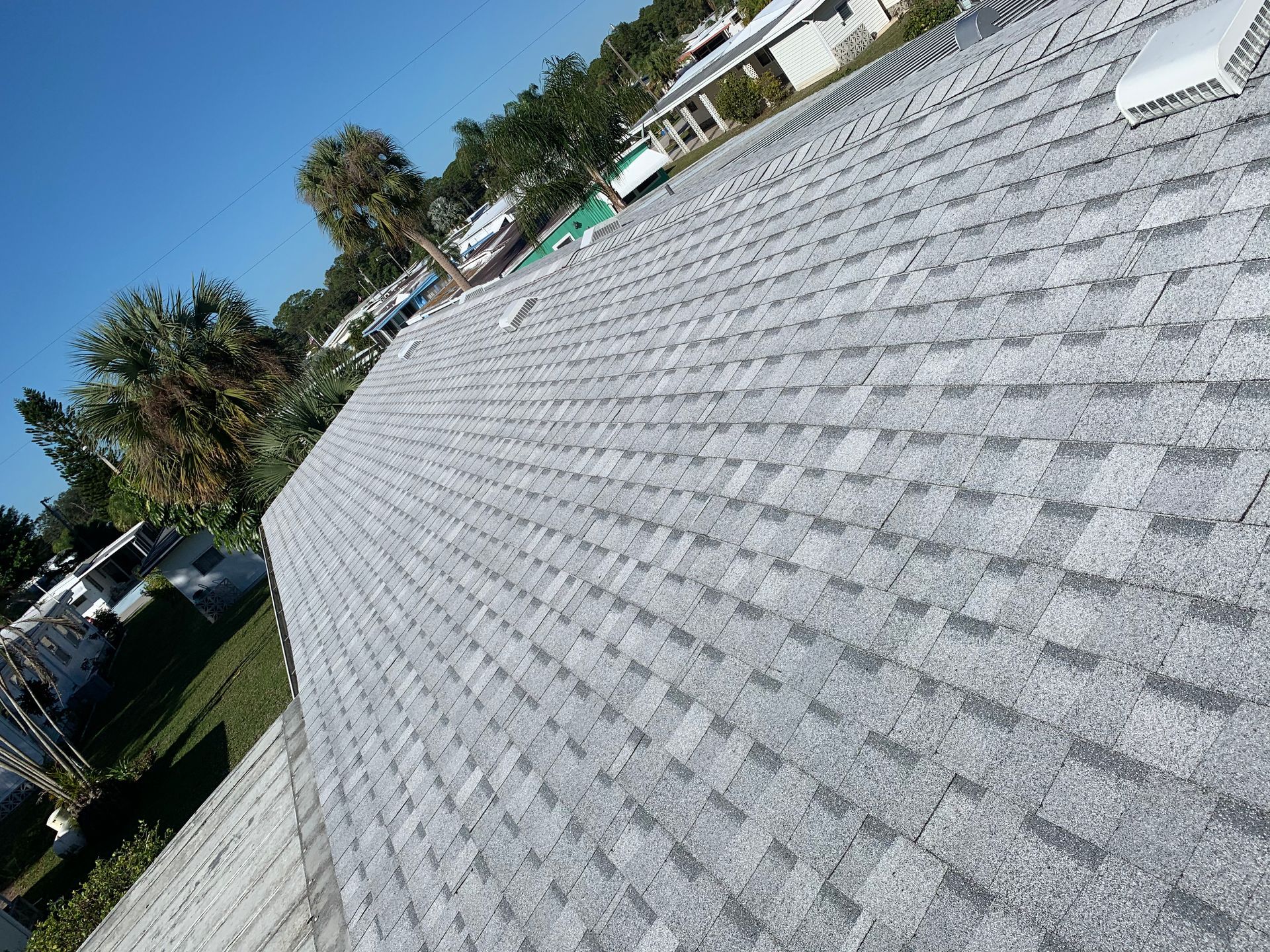 Tilted view of a gray shingled roof with palm trees and houses along the left edge under a clear sky