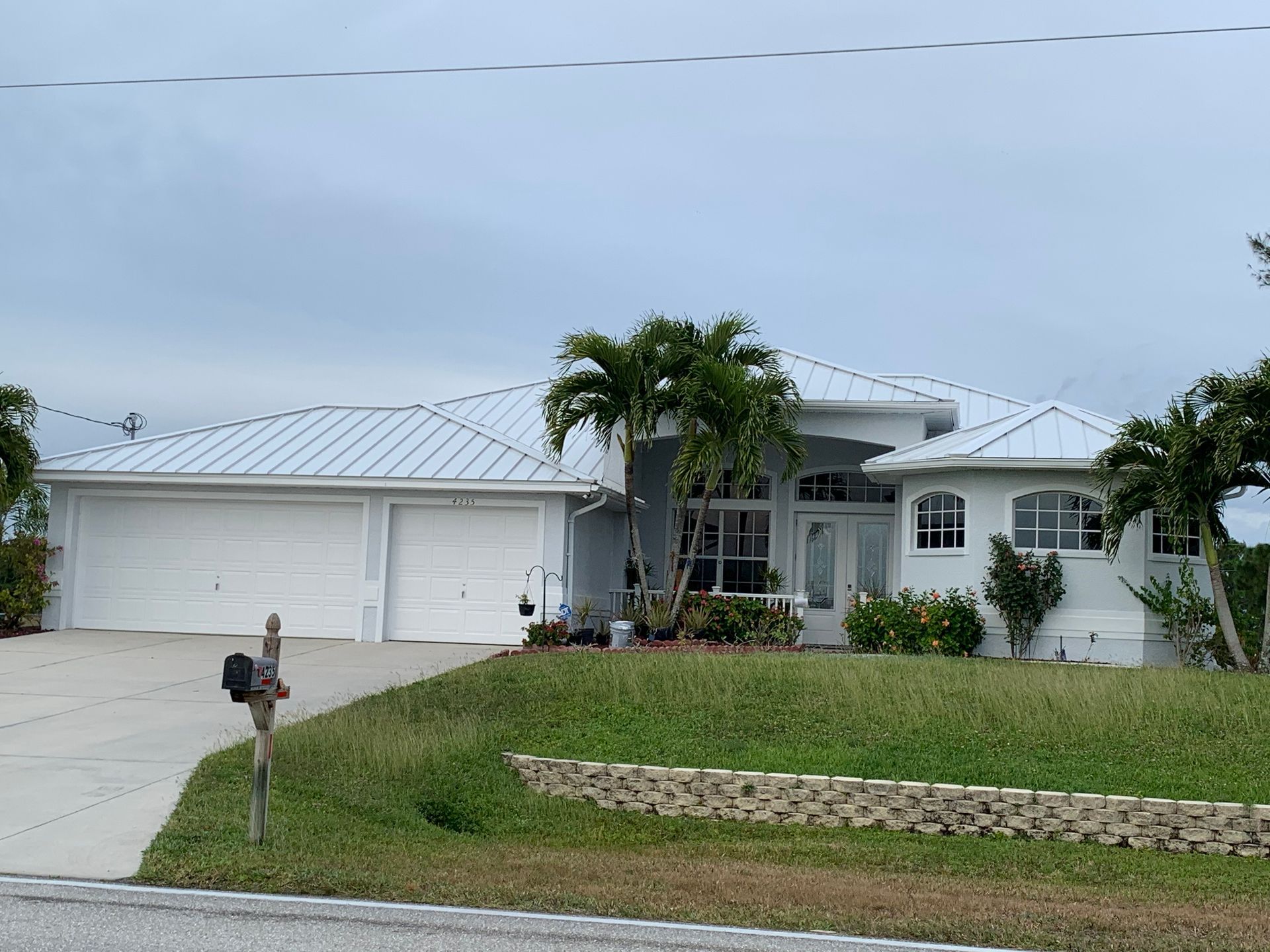 White single-story house with a metal roof, driveway, palm trees, and a front lawn by a stone retaining wall