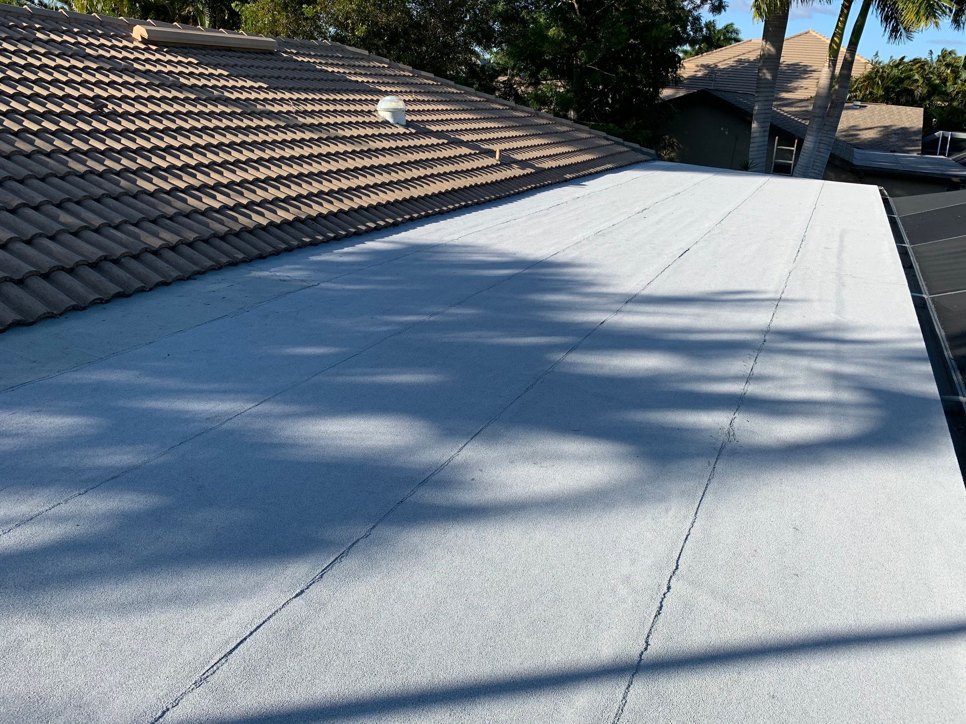 Flat rooftop with white coating next to brown tiled roof, trees casting shadows