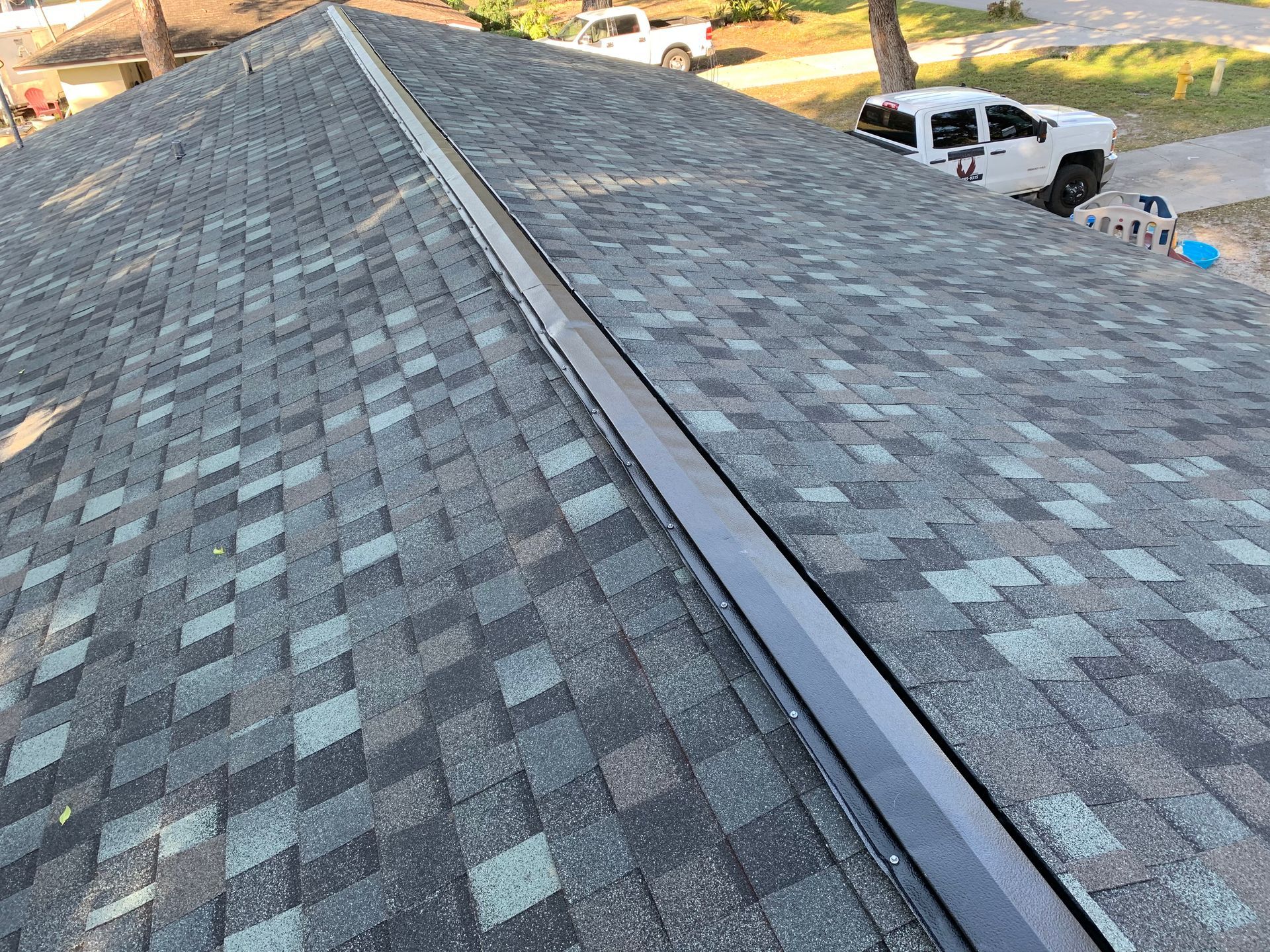 Roof with gray shingles and a central ridge vent, viewed from above with a driveway and parked trucks below