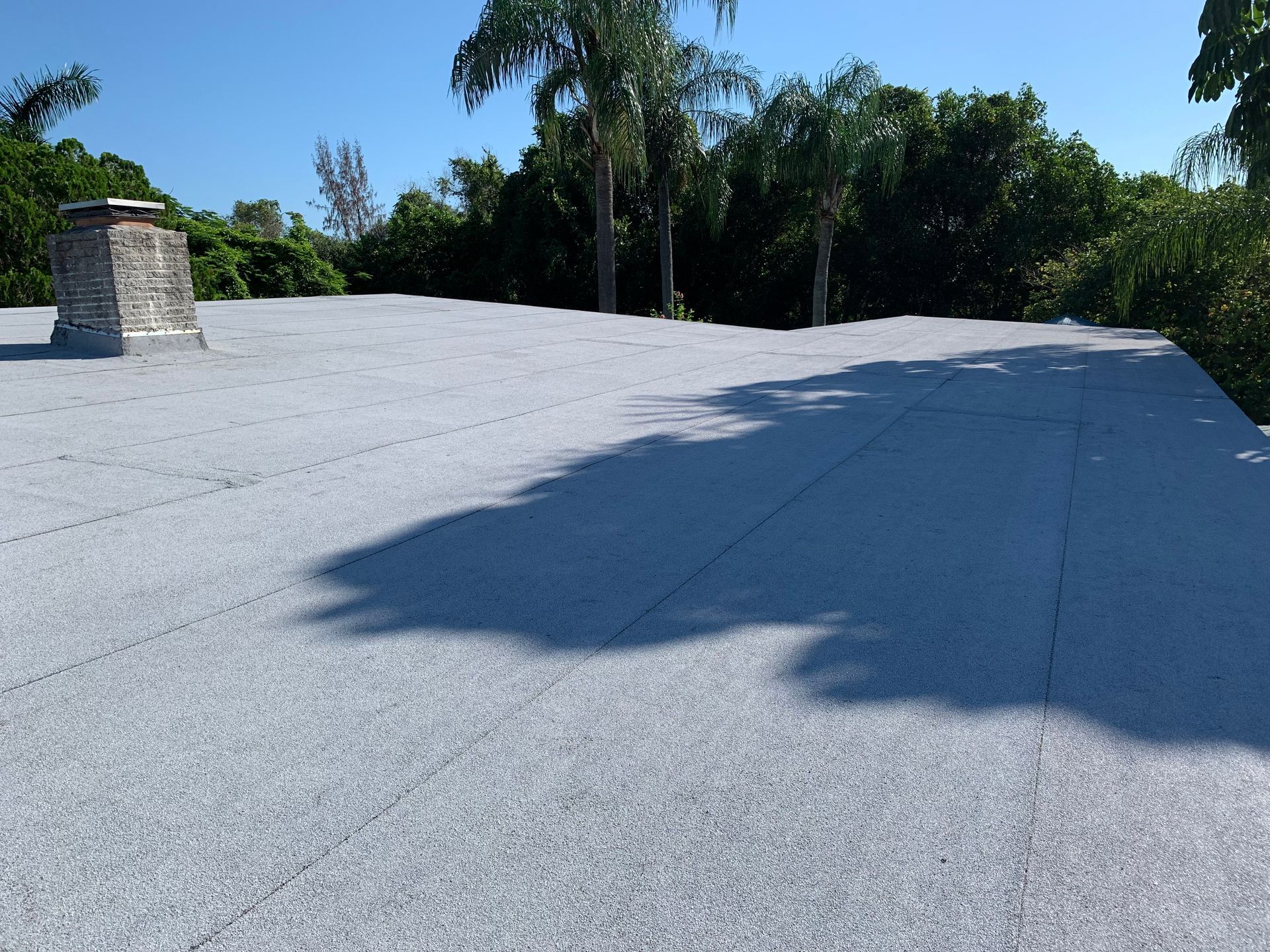 Flat white rooftop with tree shadows beside a stone chimney under a clear blue sky