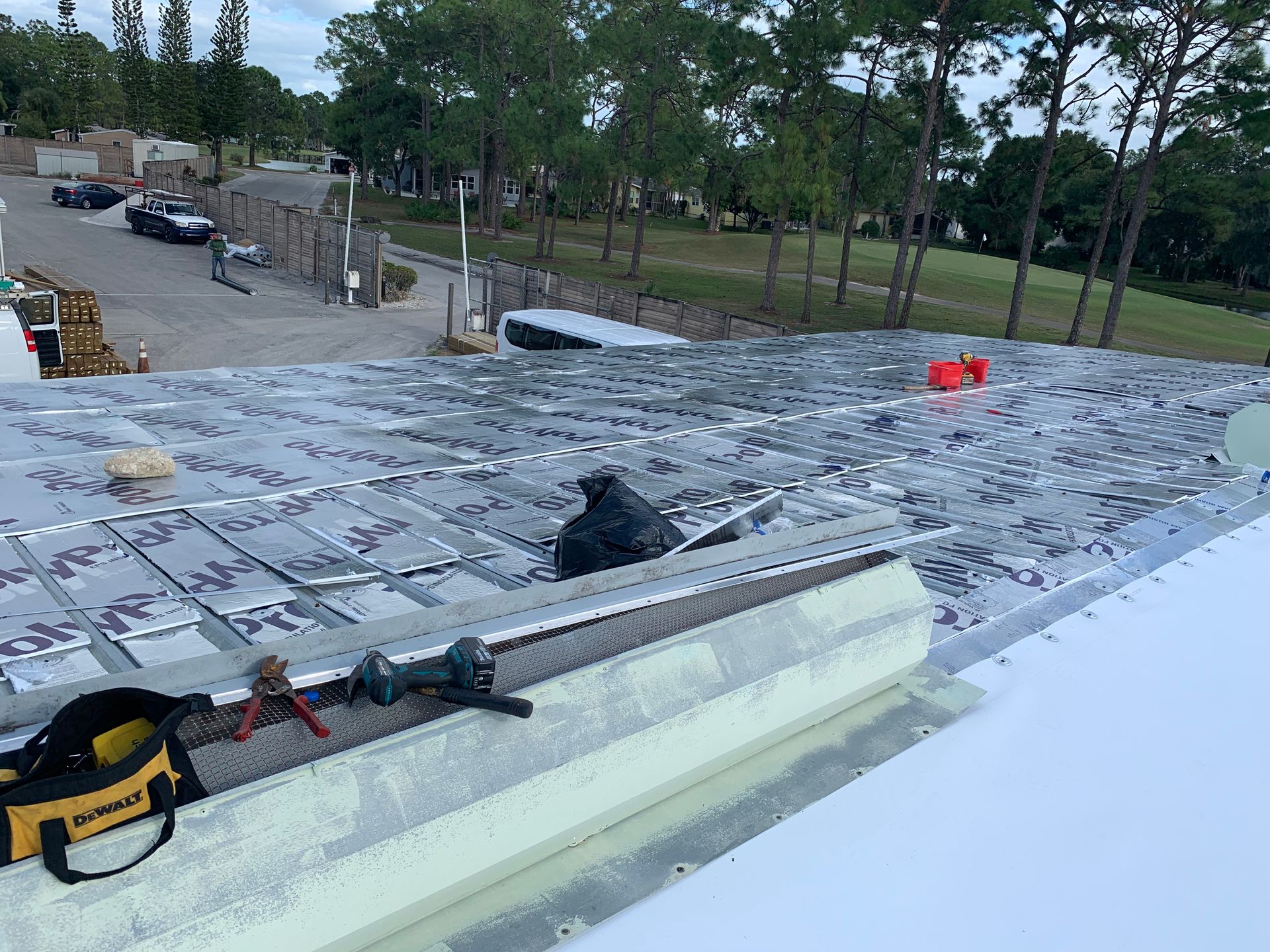 Rooftop under construction with tools, insulation, and workers in the distance surrounded by trees