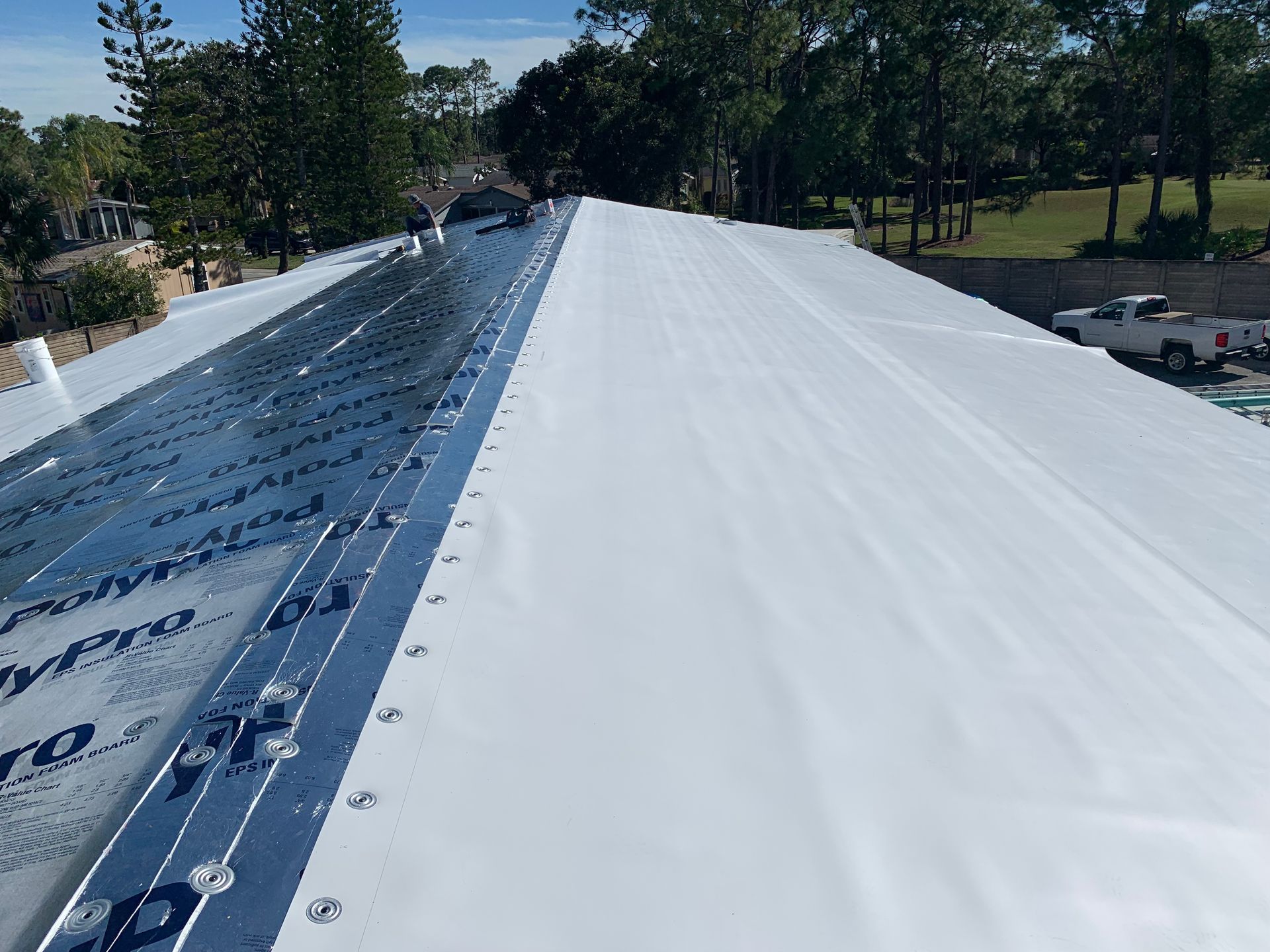 Rooftop under construction with white membrane and metal seams stretching into a tree-lined area.