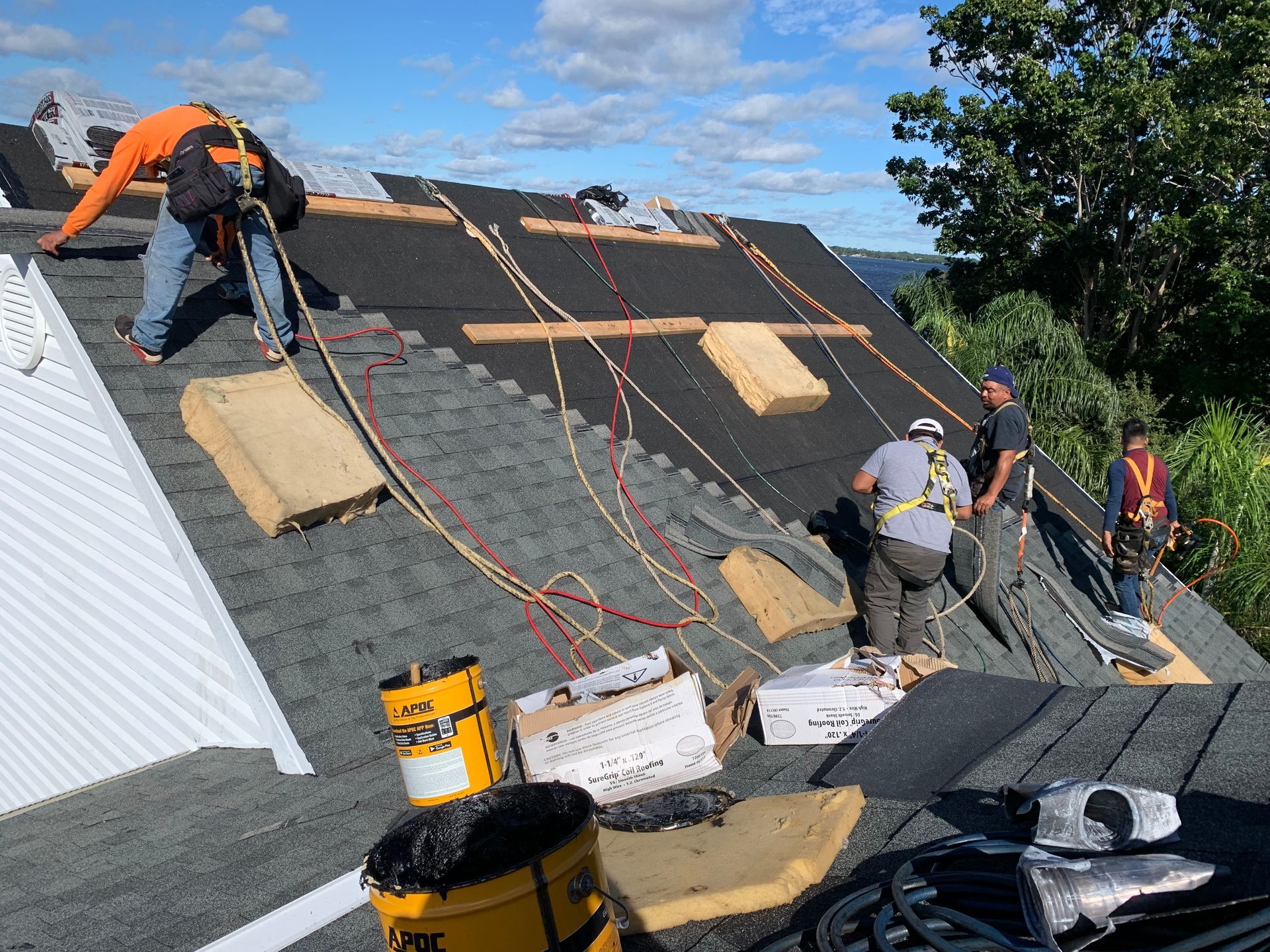 Roofers installing underlayment on a steep roof, with ropes and tools, under a blue sky.