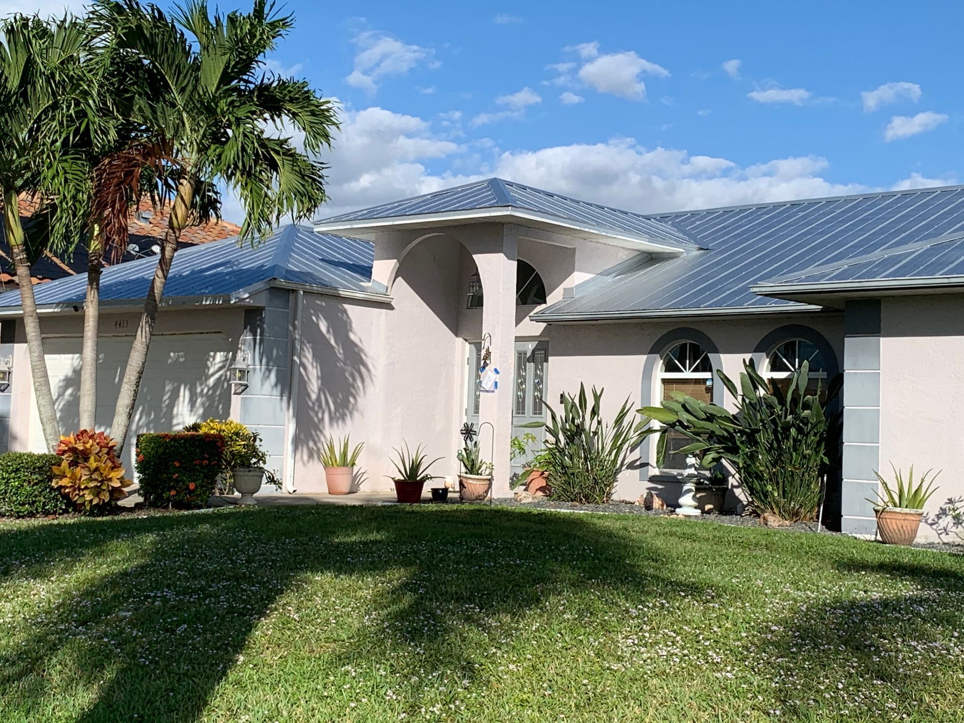 Single-story suburban house with blue roof, palm trees, and a green front lawn under a sunny sky