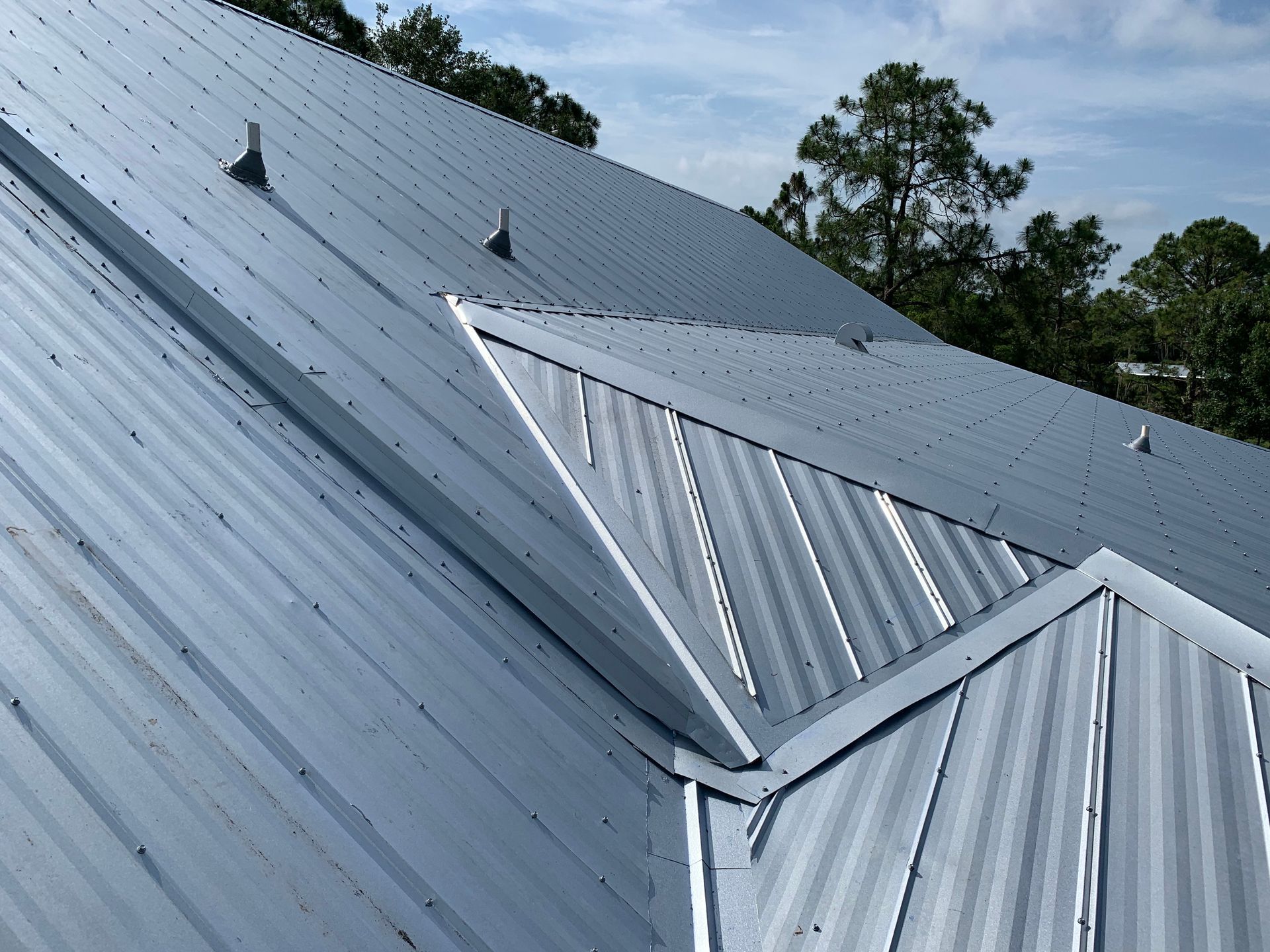 Metal roof with several ridges and vents on a sloped building, with trees in the background