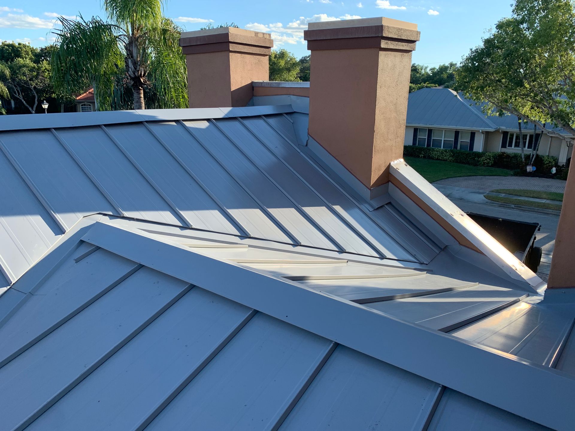 Gray metal roof with two tan chimneys in a suburban neighborhood