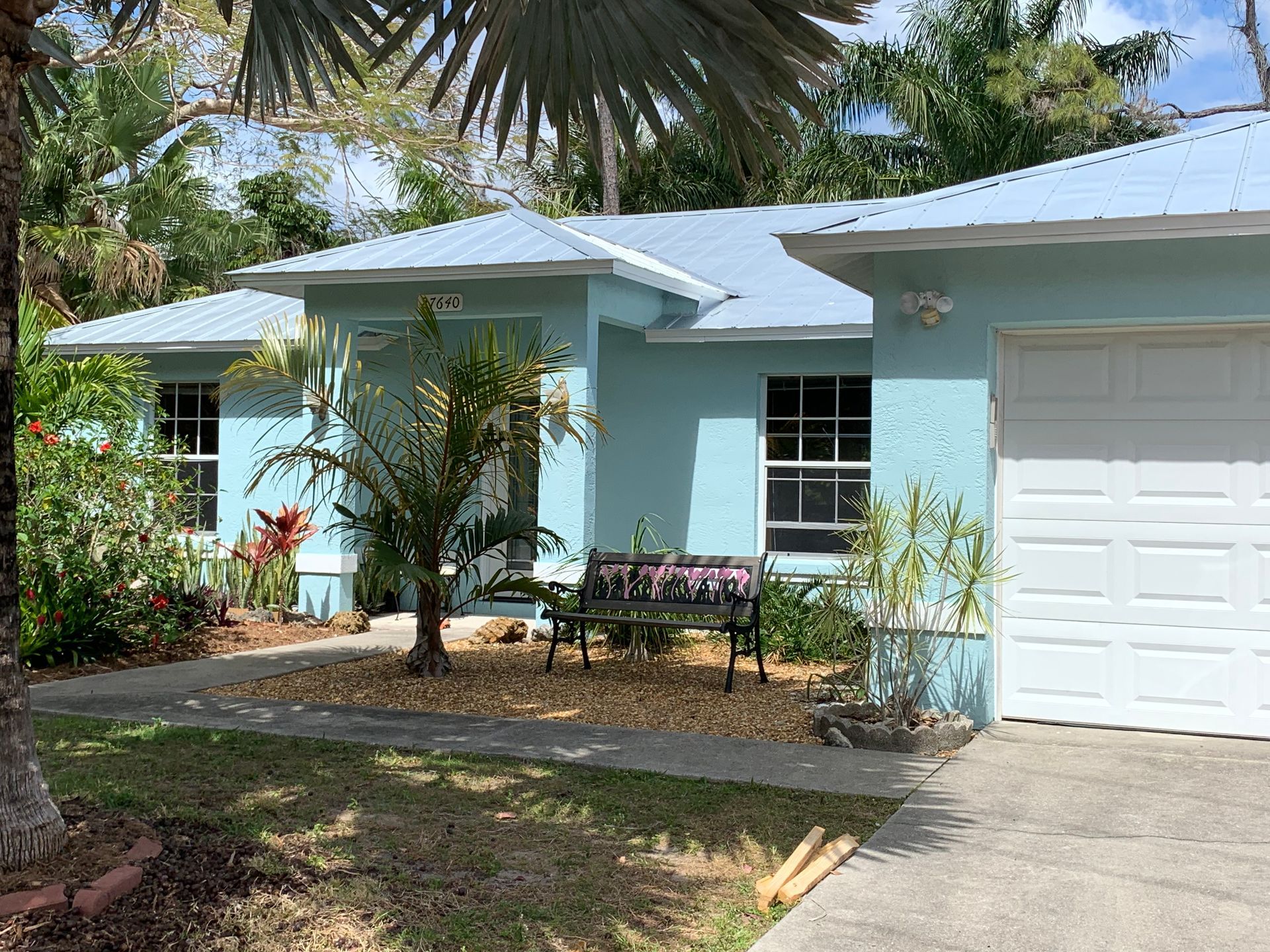Light blue house with a garage, front garden, and palm trees in a sunny yard.