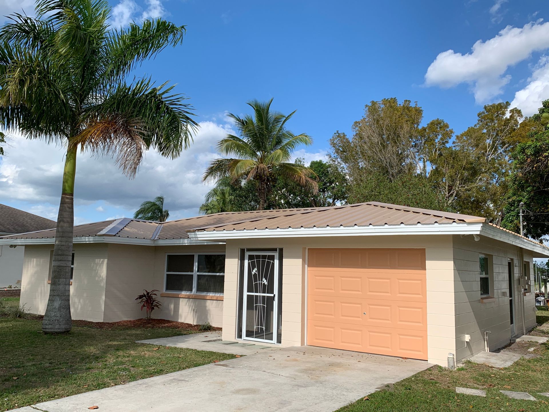 Single-story suburban house with peach garage door, white exterior, driveway, palm trees, and blue sky