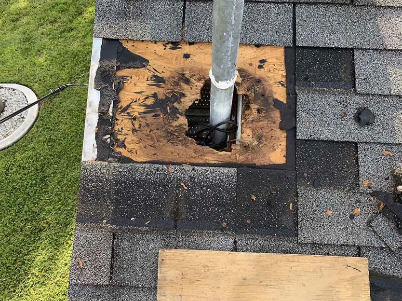 Damaged shingle roof around a vent pipe, with exposed plywood and a patched board nearby