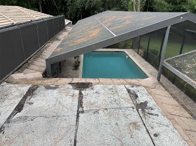 Outdoor pool covered by a sloped gray roof, seen from above with patio paving and glass fencing.