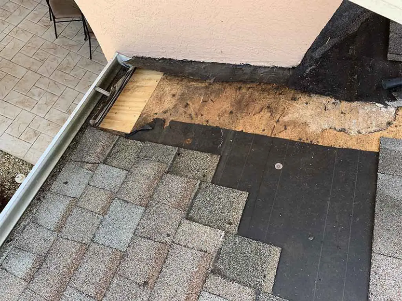Damaged shingle roof around a vent pipe, with exposed plywood and a patched board nearby