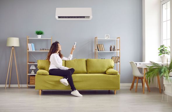 Woman sitting on a lime-green sofa in a bright living room, holding up a remote toward a wall-mounted air conditioner