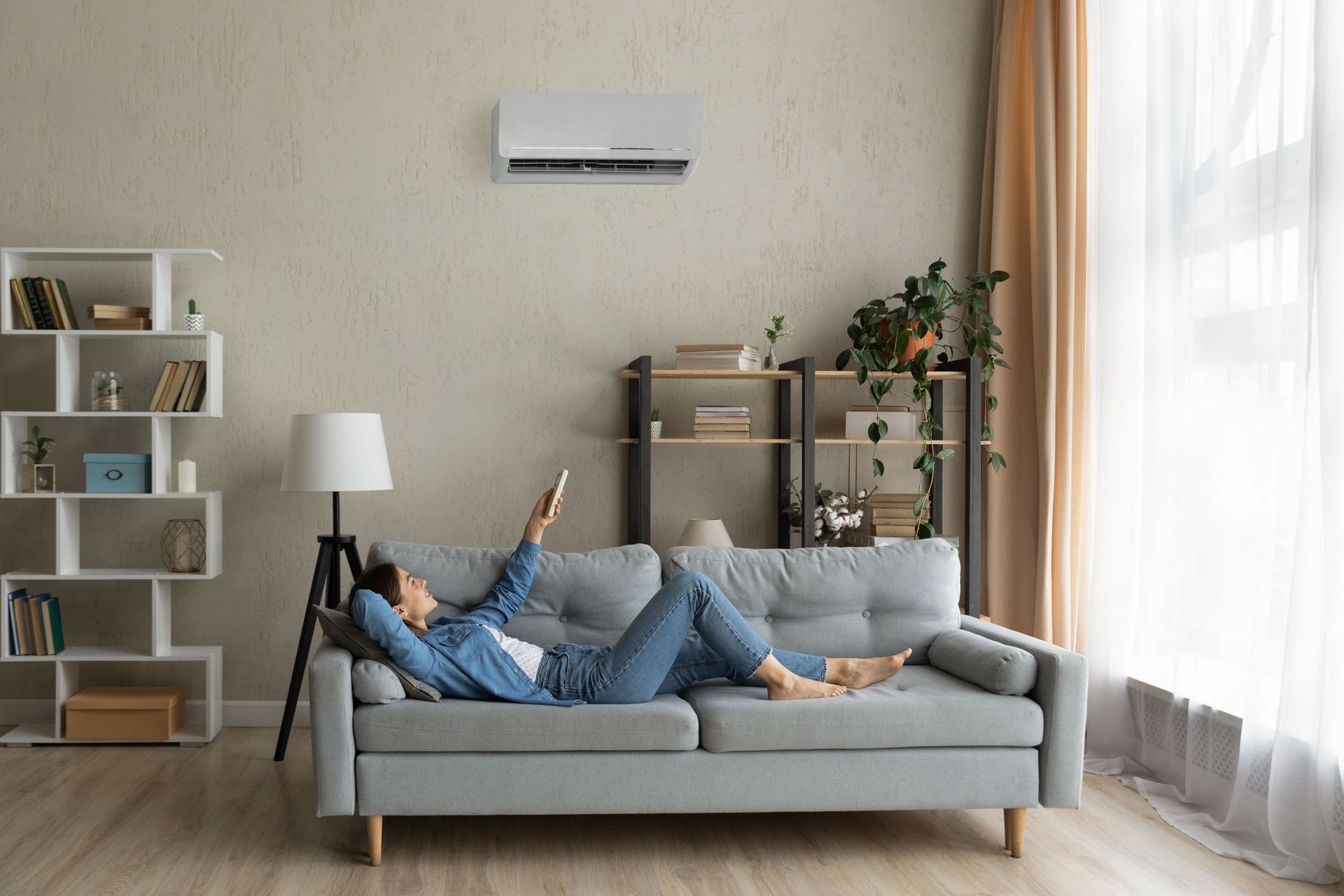 Person relaxing on a gray sofa in a bright, minimalist living room with shelves, lamp, and plants