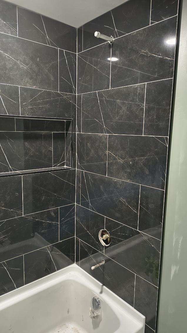 Small bathroom shower with black tiled walls, white tub edge, and a wall-mounted showerhead.