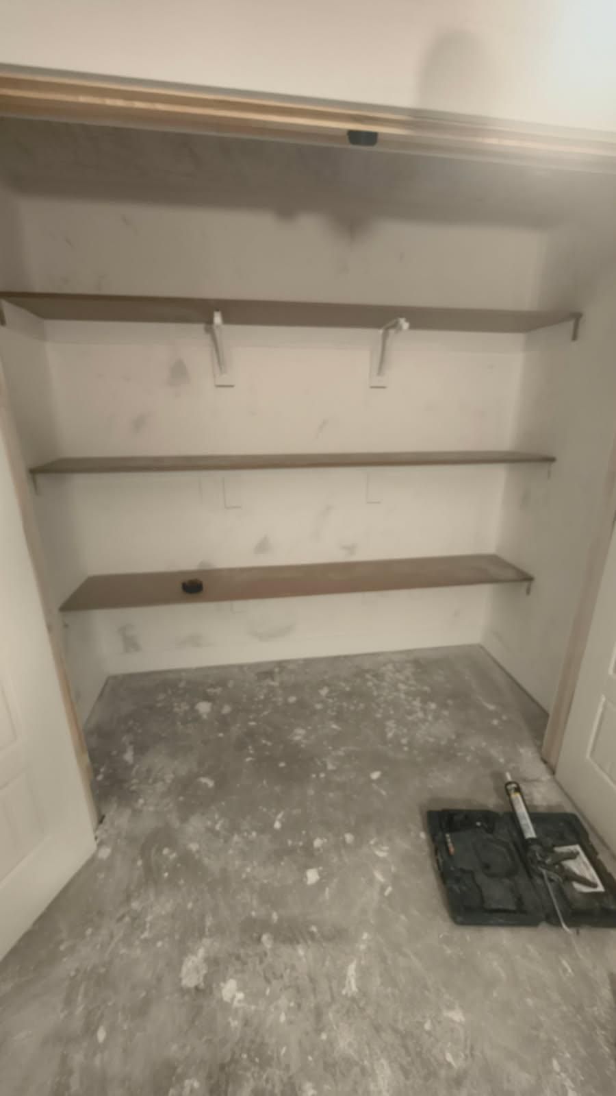 Empty closet with two wooden shelves, hanging rods, and a bare floor with debris and tools.