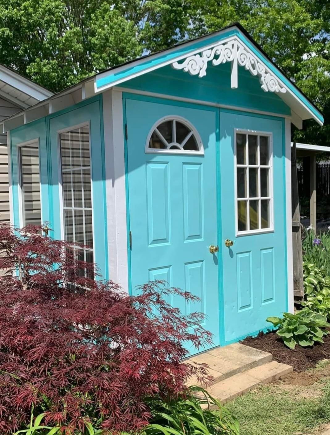 A bright teal garden shed with white trim, a decorative gable, double doors with windows, and leafy plants in front