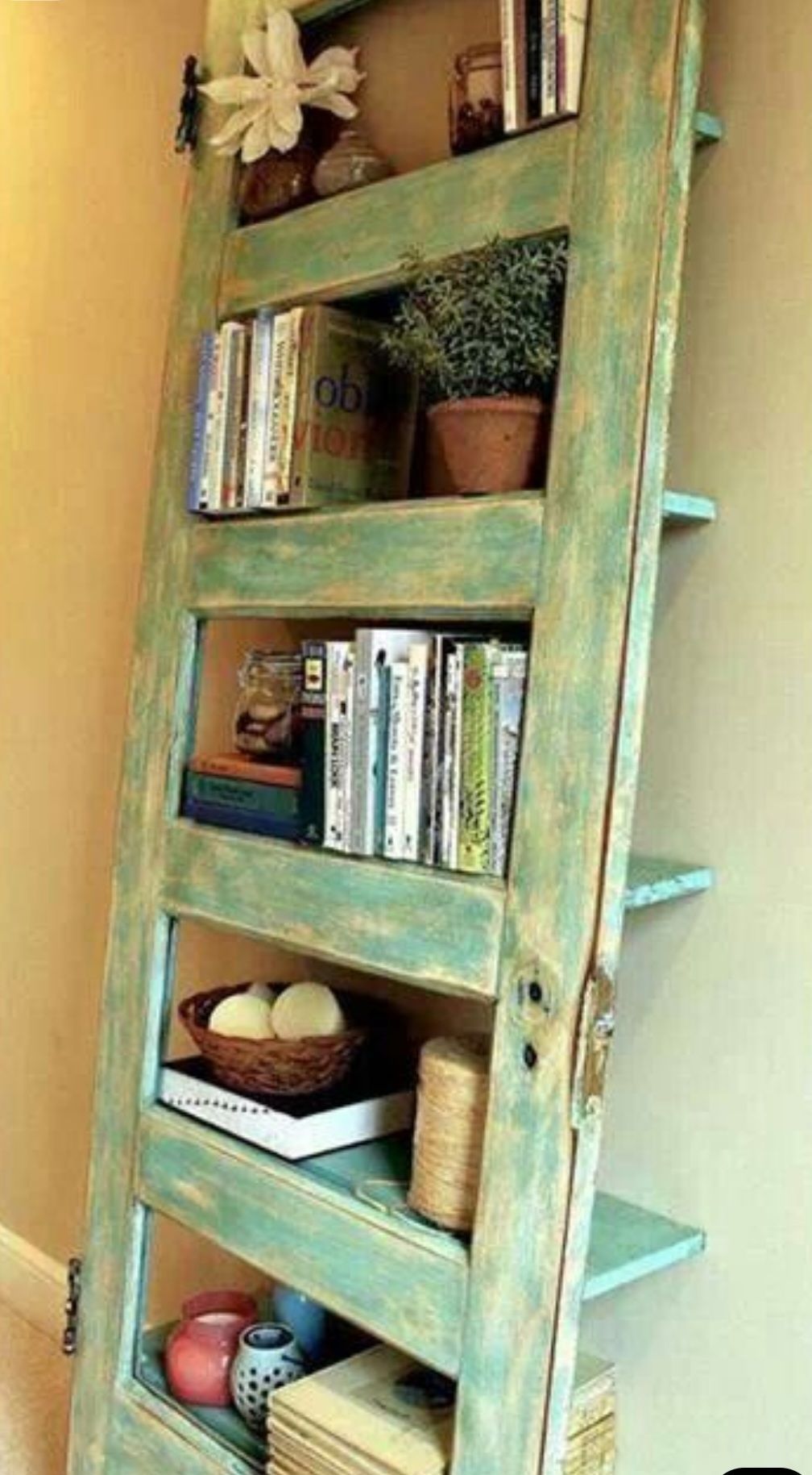 A distressed mint-green painted door repurposed into a leaning bookshelf against a beige wall, holding books and decor