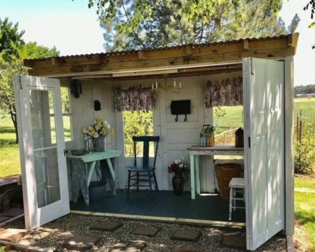 An open-sided, rustic shed set outdoors, furnished with tables, a wooden chair, floral curtains, and a gravel floor
