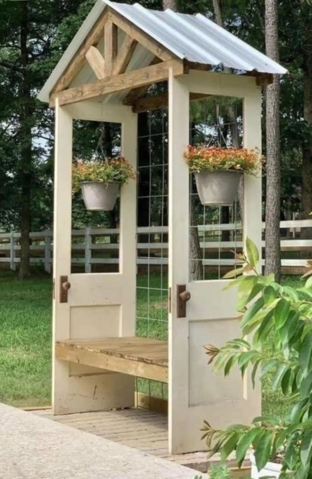 A garden bench built from two white doors with a metal roof and hanging flower pots, set on a patio in a backyard