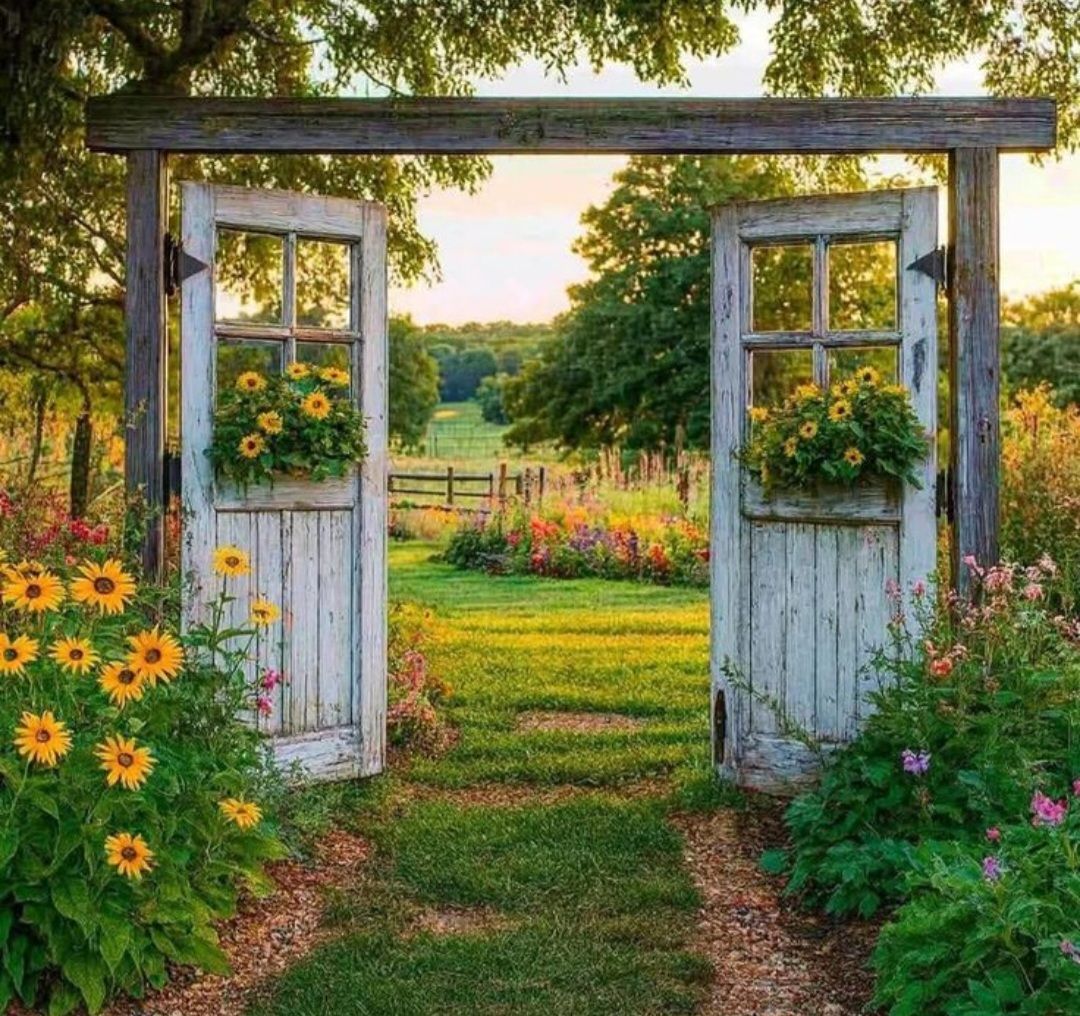Two weathered white doors set in a wooden frame serve as a garden gate, surrounded by sunflowers and green fields at sunset