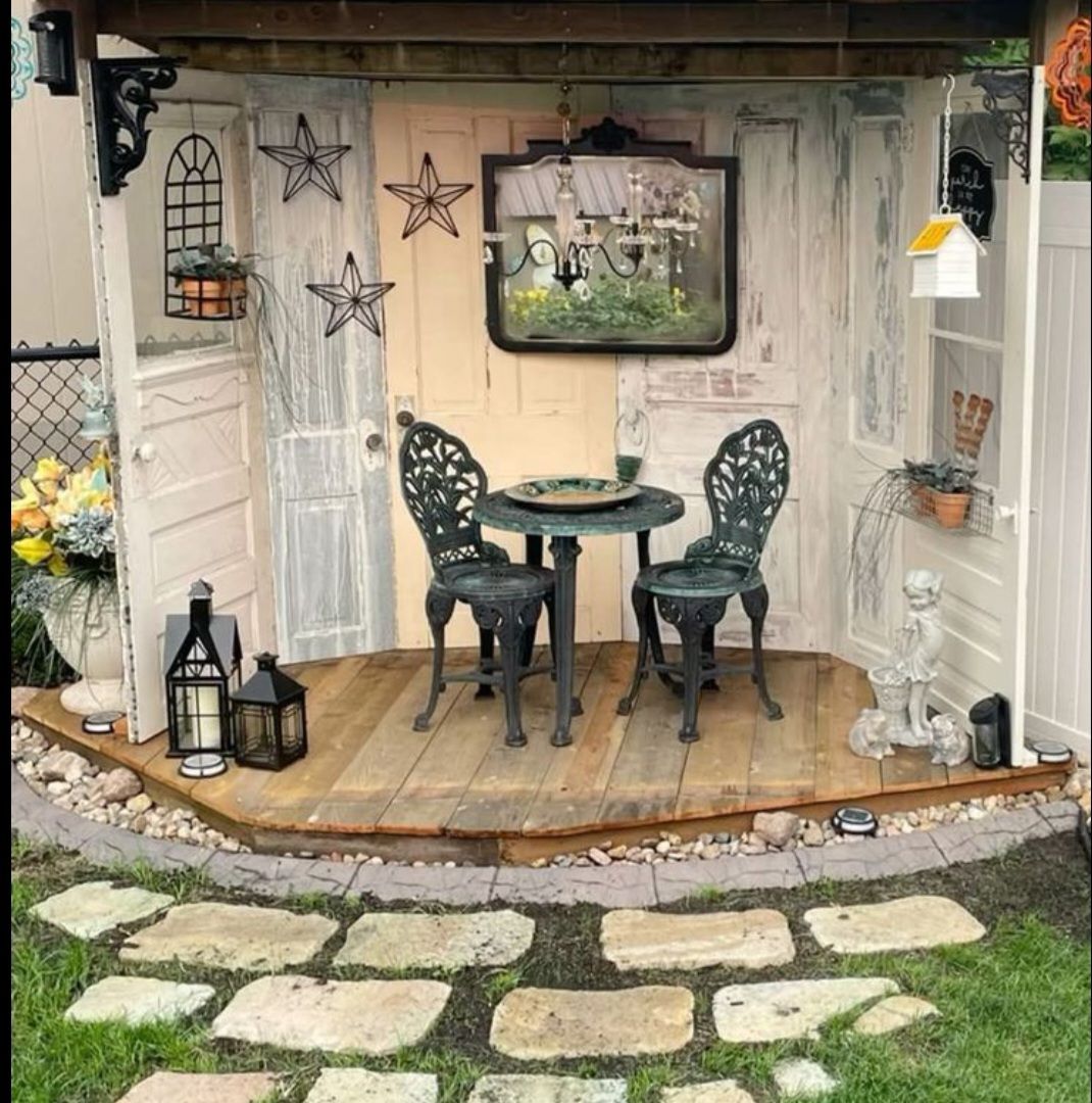 A small, rustic outdoor bistro set with two dark metal chairs and a table on a wooden deck framed by vintage white doors