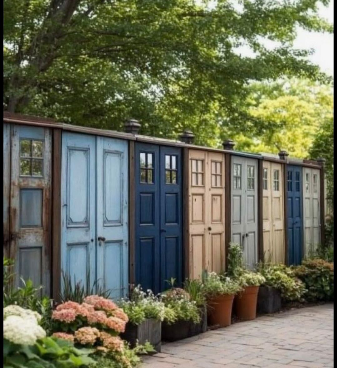 A backyard fence constructed from colorful vintage wooden doors, with potted plants lining the base in front