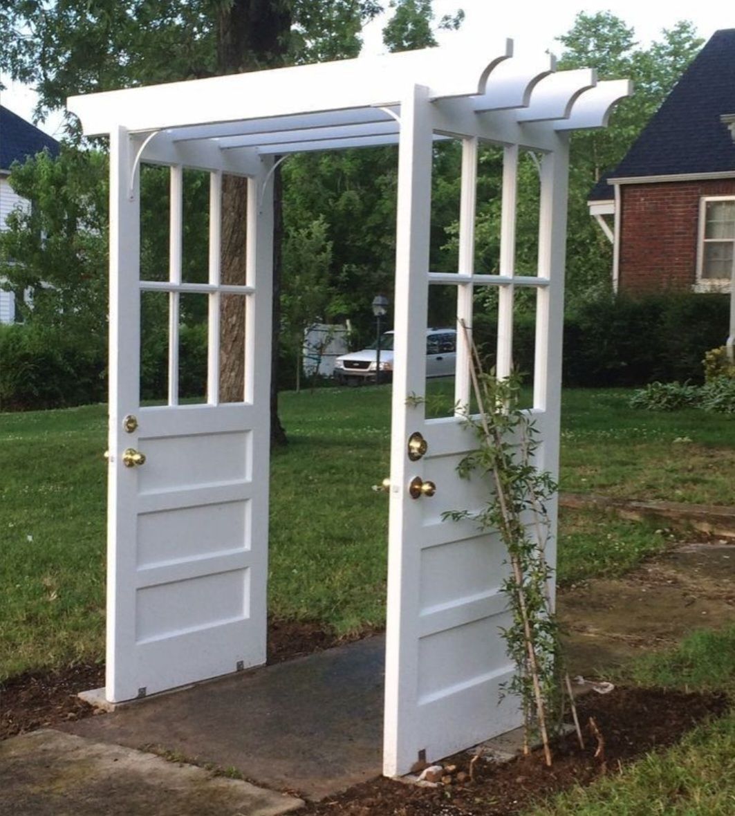 A white garden arbor made from two repurposed vintage doors with glass panels, standing over a path in a grassy yard