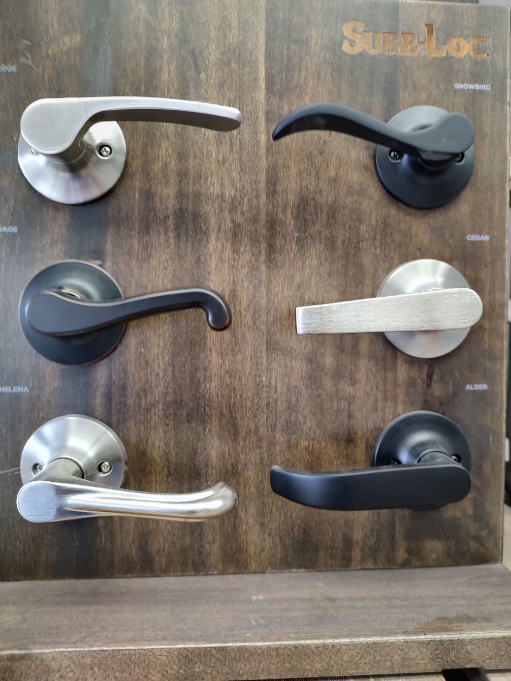 A wooden display board featuring six different styles and finishes of metal door lever handles arranged in two columns.