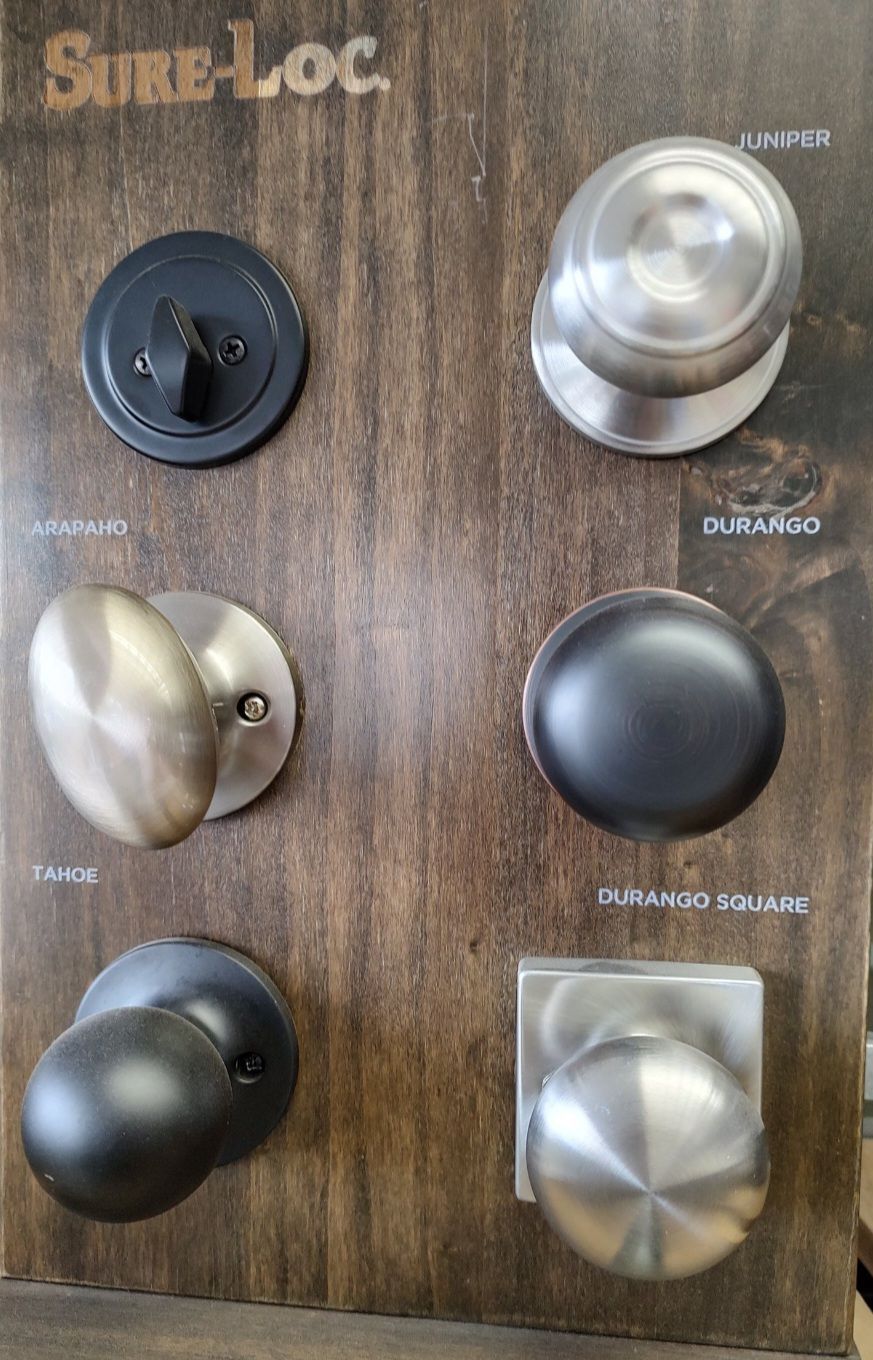 A display board from Sure-Loc showing six different metal door knob and deadbolt styles in matte black and silver finishes.