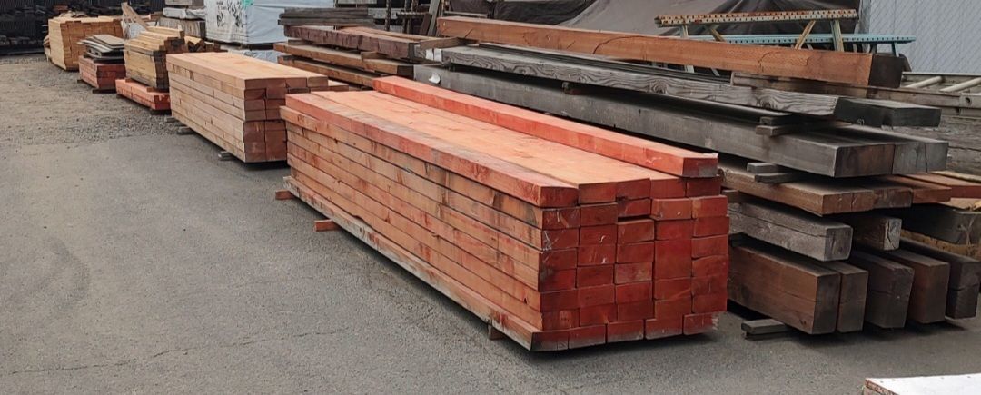 Piles of assorted wooden beams and construction lumber stacked on a paved outdoor lot