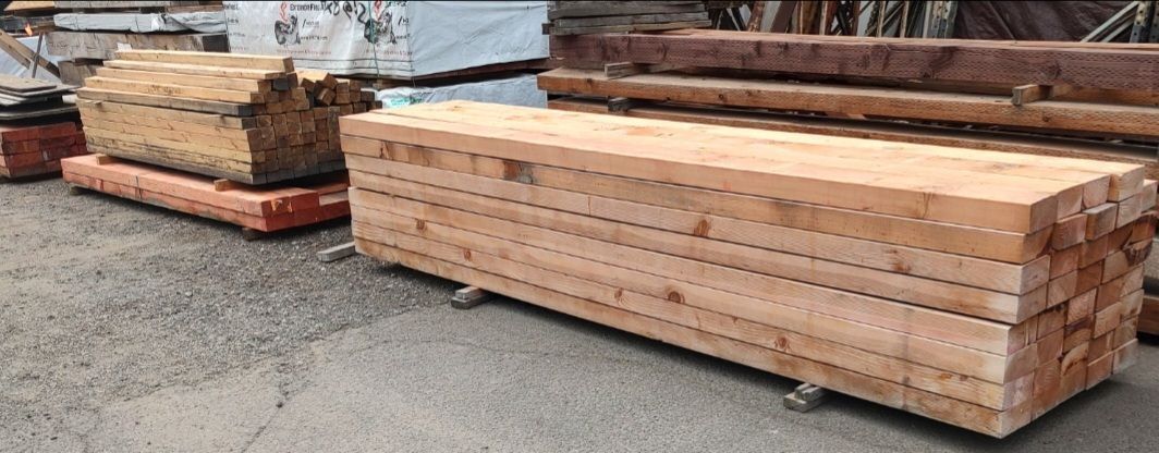 Stacked bundles of raw, light-colored wooden beams resting on a paved outdoor lot