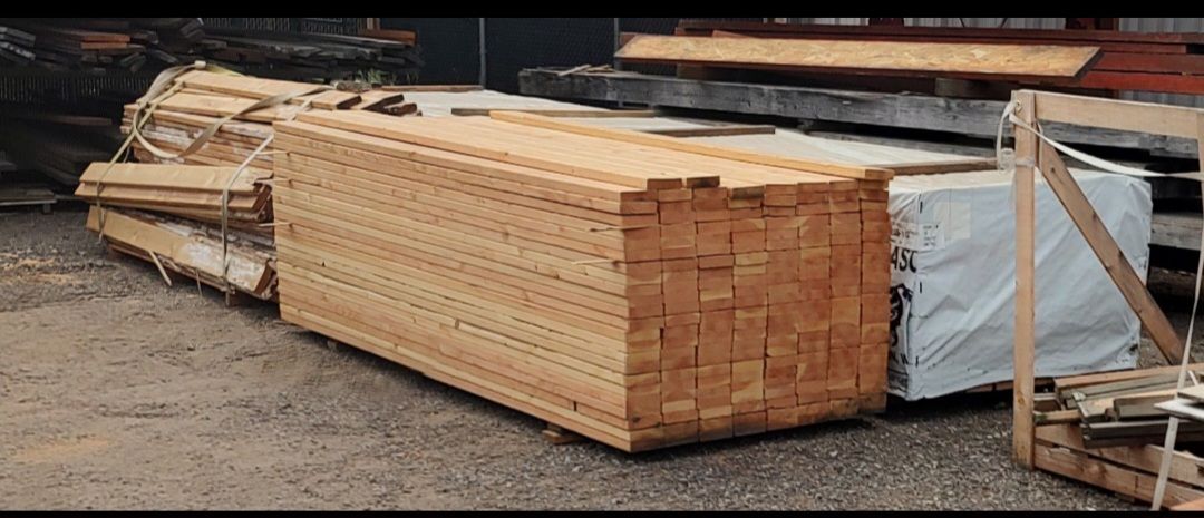 Stacked lumber of various sizes outdoors, including bundles of finished planks and raw wood