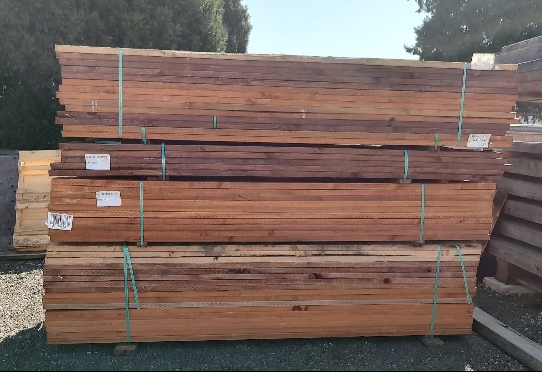 A stack of rough-cut wooden planks with visible grain, banded with green plastic straps and stored on a gravel lot