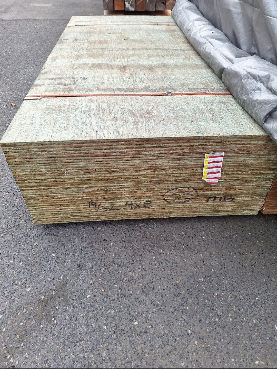 A stack of plywood sheets stored outdoors on a paved surface, partially covered by a gray tarp