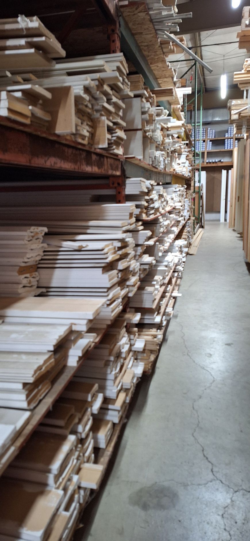 A warehouse aisle with tall shelves packed with stacked wooden boards, moldings, and trim pieces