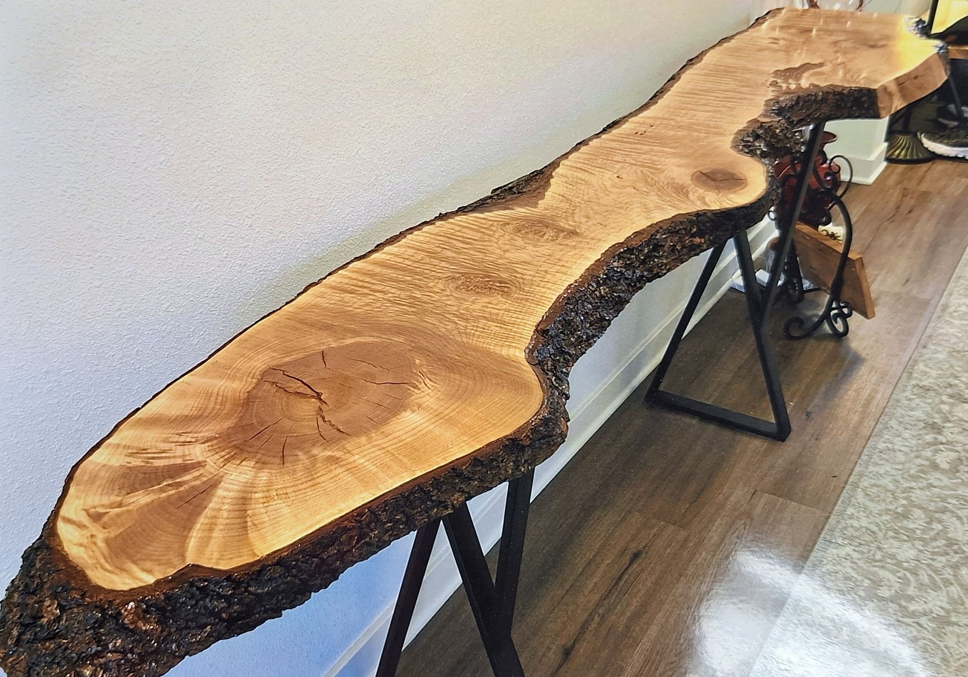 A live-edge wood console table with a natural bark finish and metal legs stands against a wall on a wood-look floor