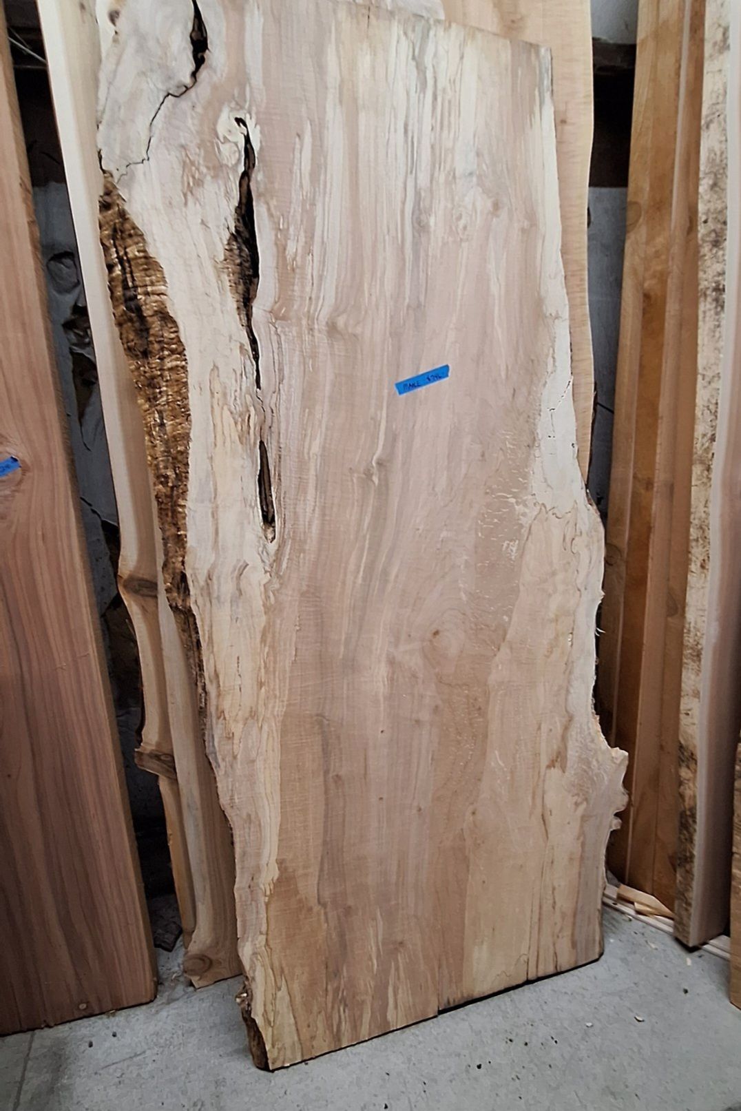 A large, vertical raw wood slab with a rough, natural bark edge and a deep, vertical split standing in a lumber yard
