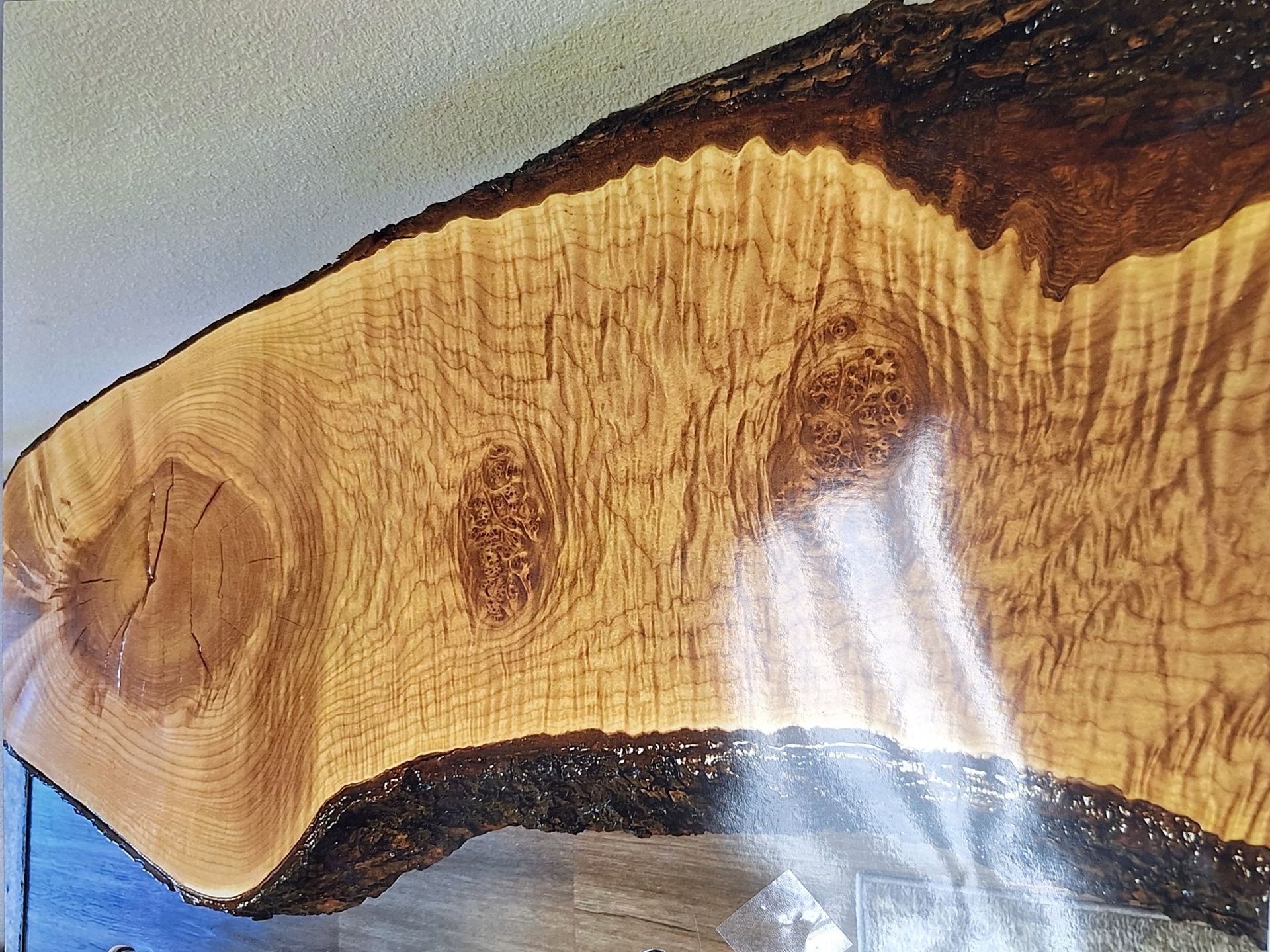 A raw, live-edge wood slab featuring swirling light-brown grain patterns, visible knots, and dark, textured tree bark