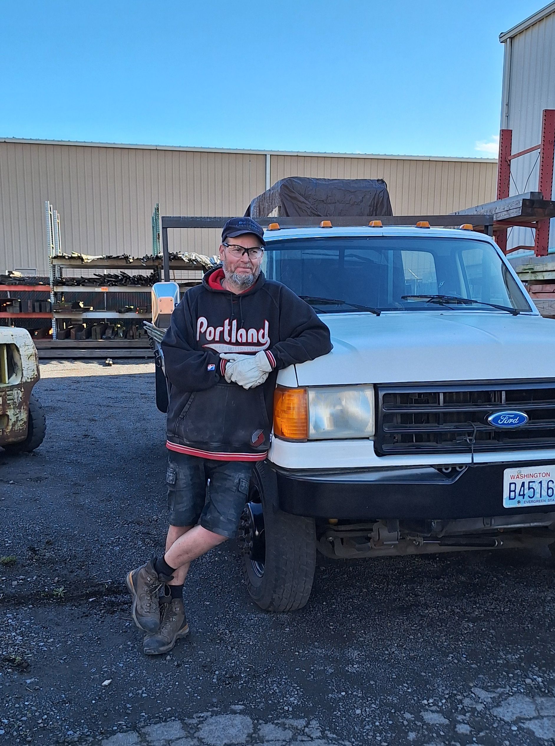 A person wearing a Portland hoodie, shorts, and work boots leans against the front of a white Ford truck in a yard.