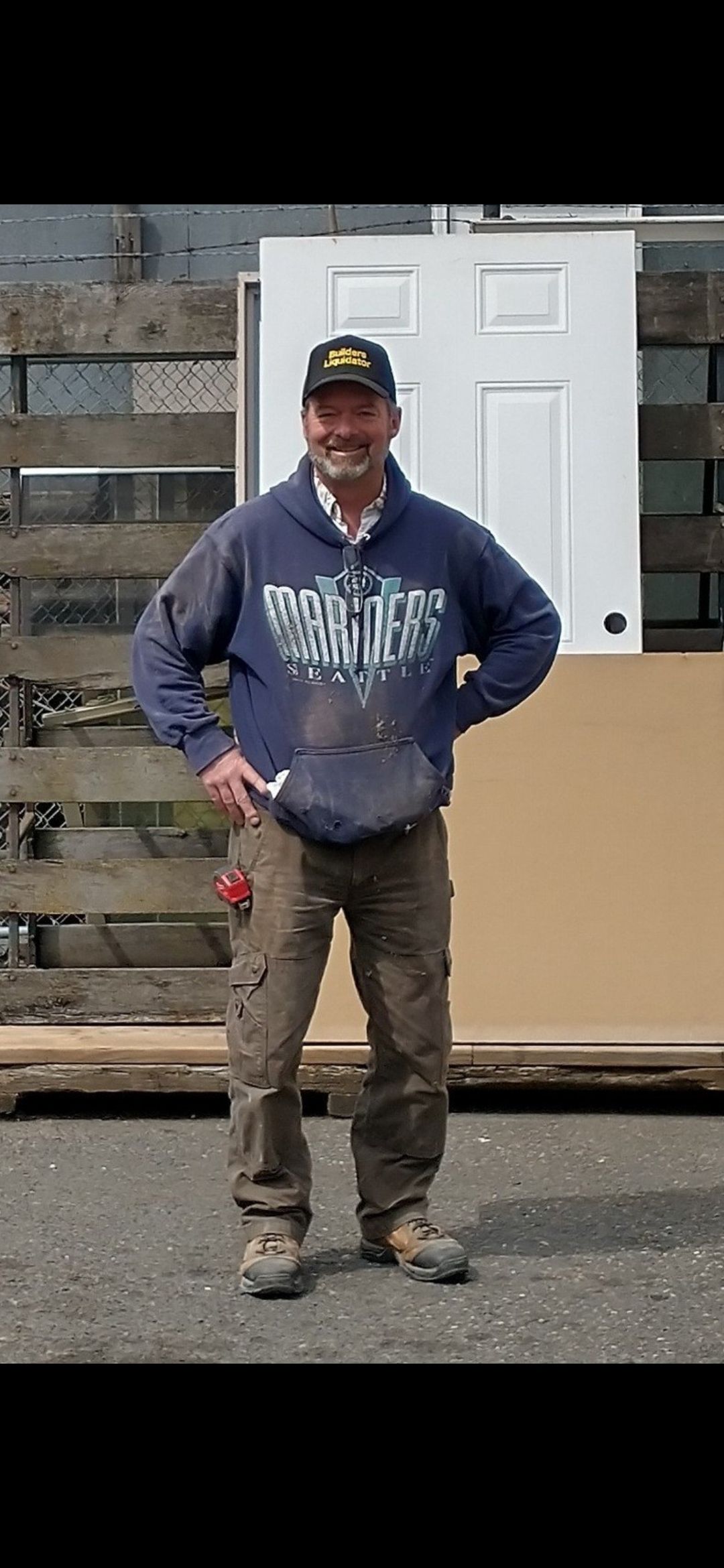 A person wearing a dark blue hoodie, brown work pants, and a baseball cap stands smiling in front of a white door frame.