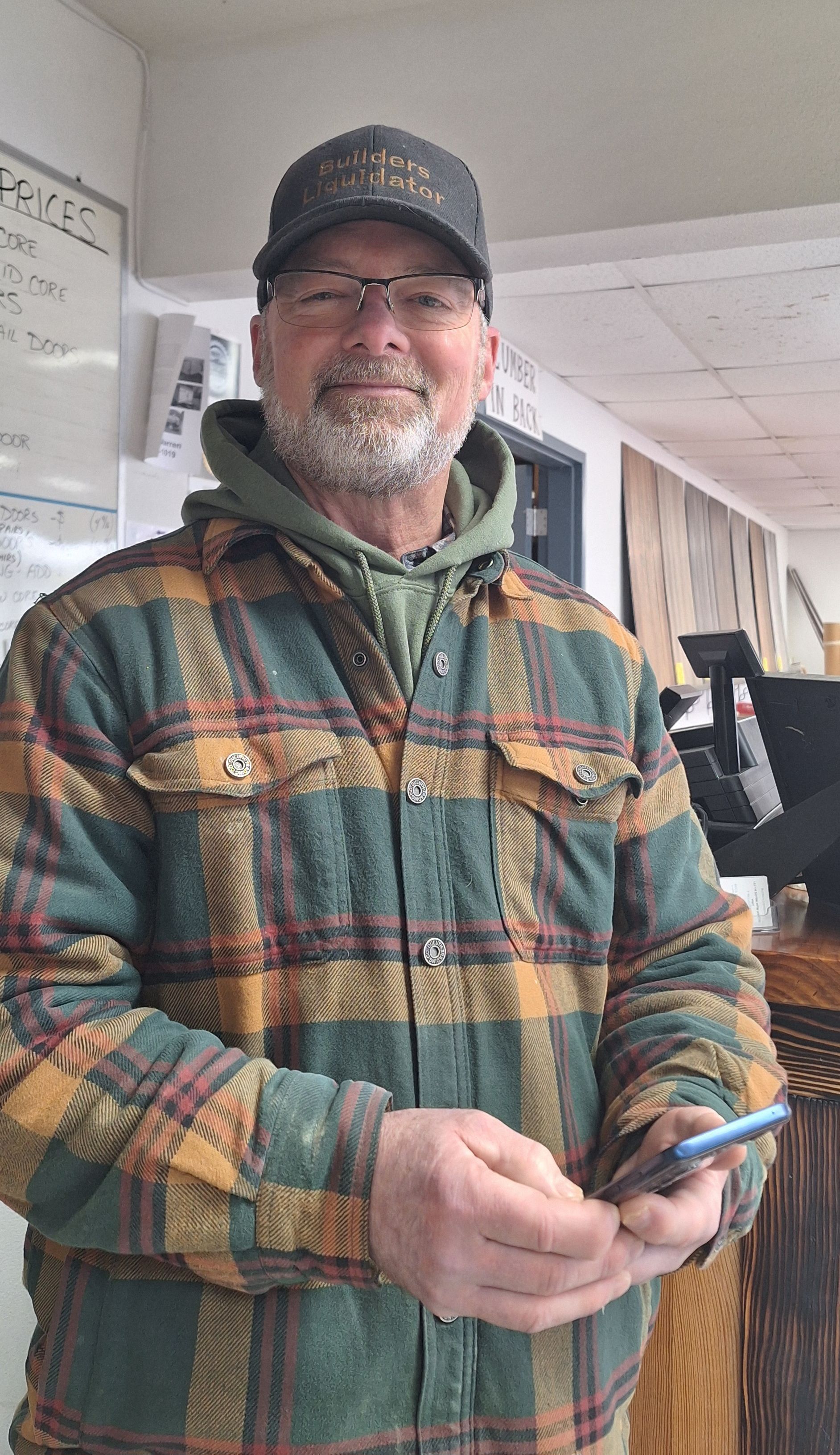 A person wearing a plaid jacket, hoodie, and cap holds a phone, standing in an indoor setting.