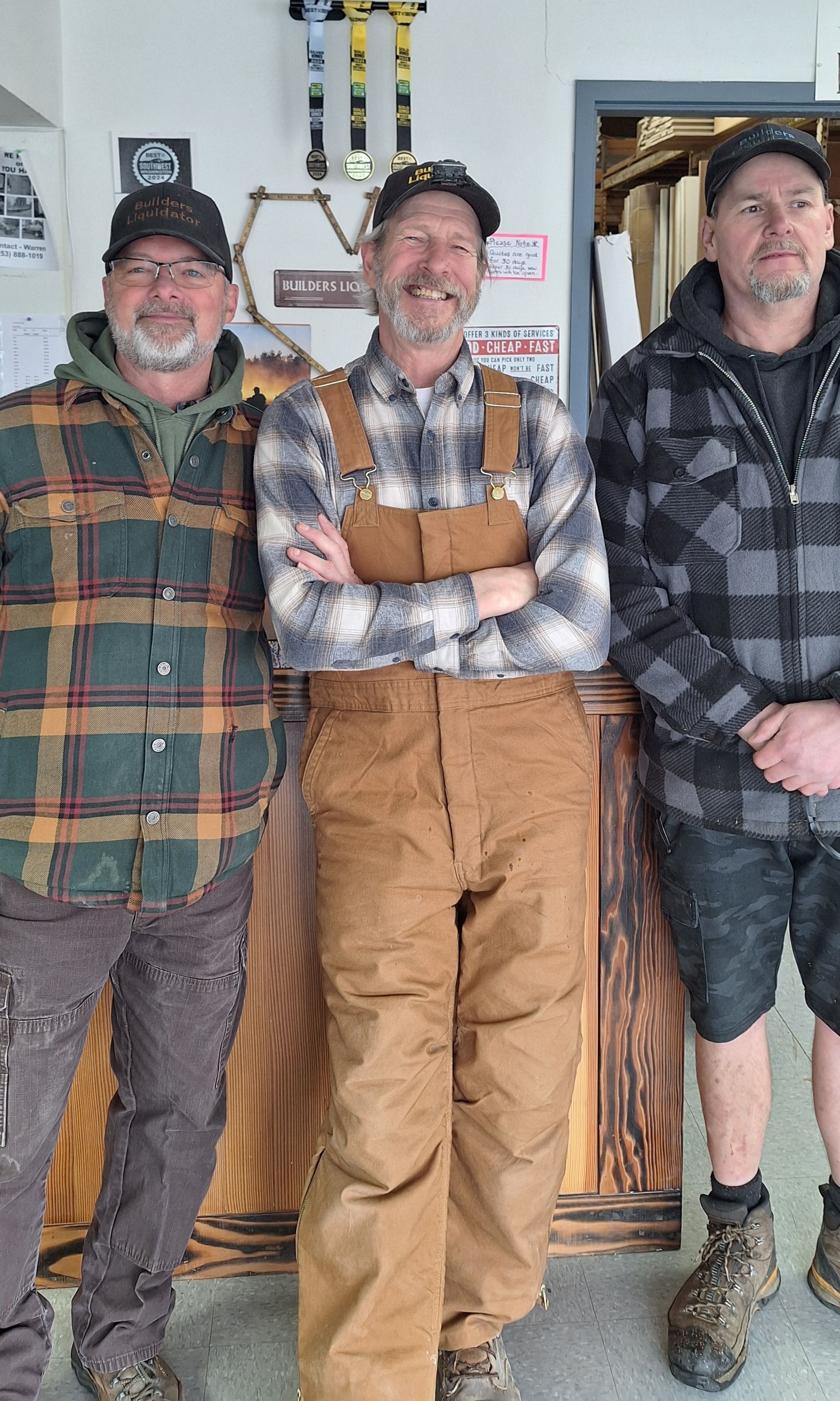 Three people wearing casual outdoor clothing stand side-by-side in a workshop, smiling at the camera.
