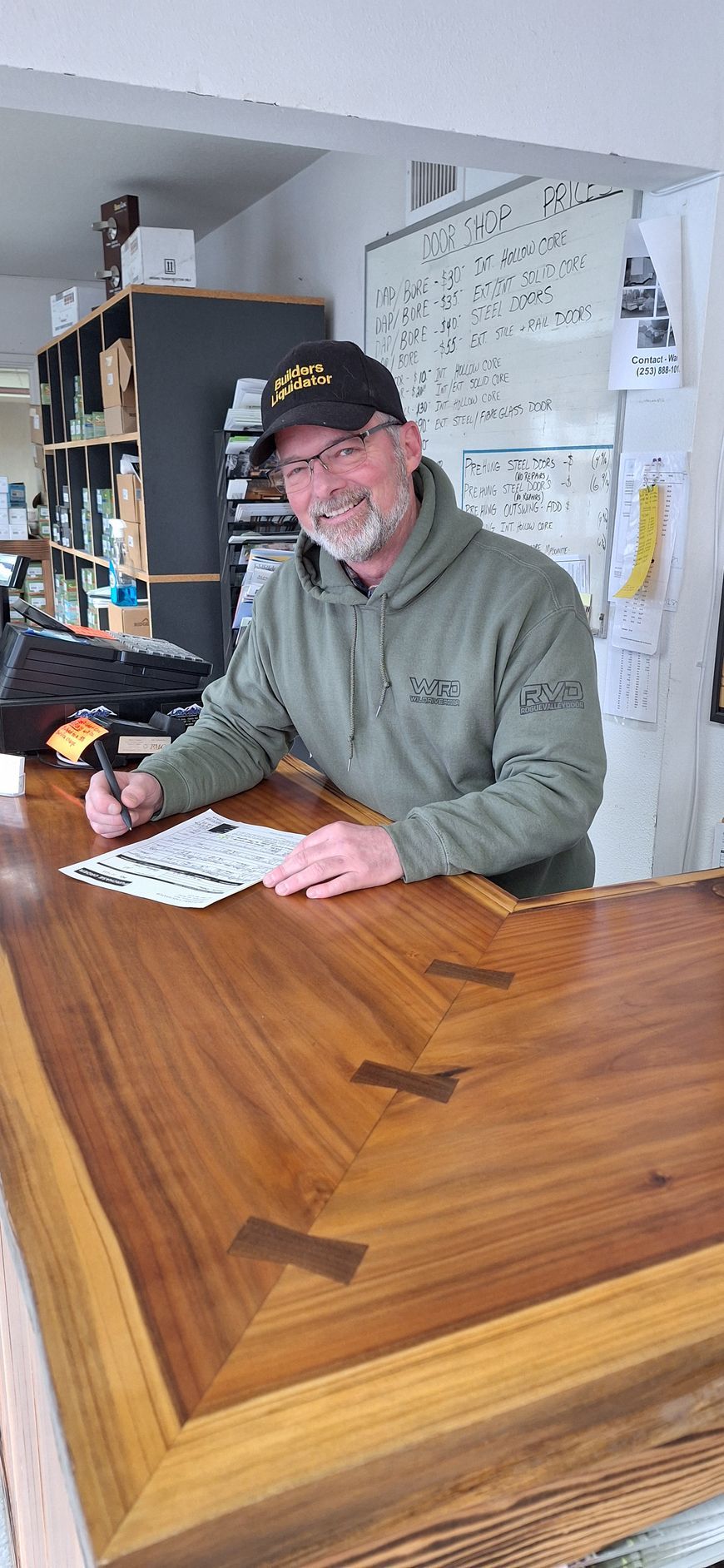 A smiling person in a green hoodie and black cap writes on a paper at a wooden counter with butterfly joint details.