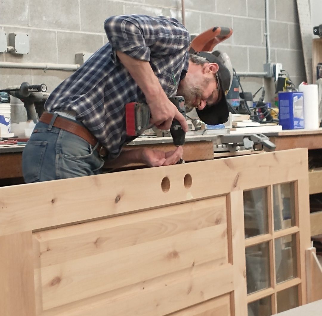 A person in a plaid shirt uses a power drill on the top edge of a wooden door frame in a woodworking shop.