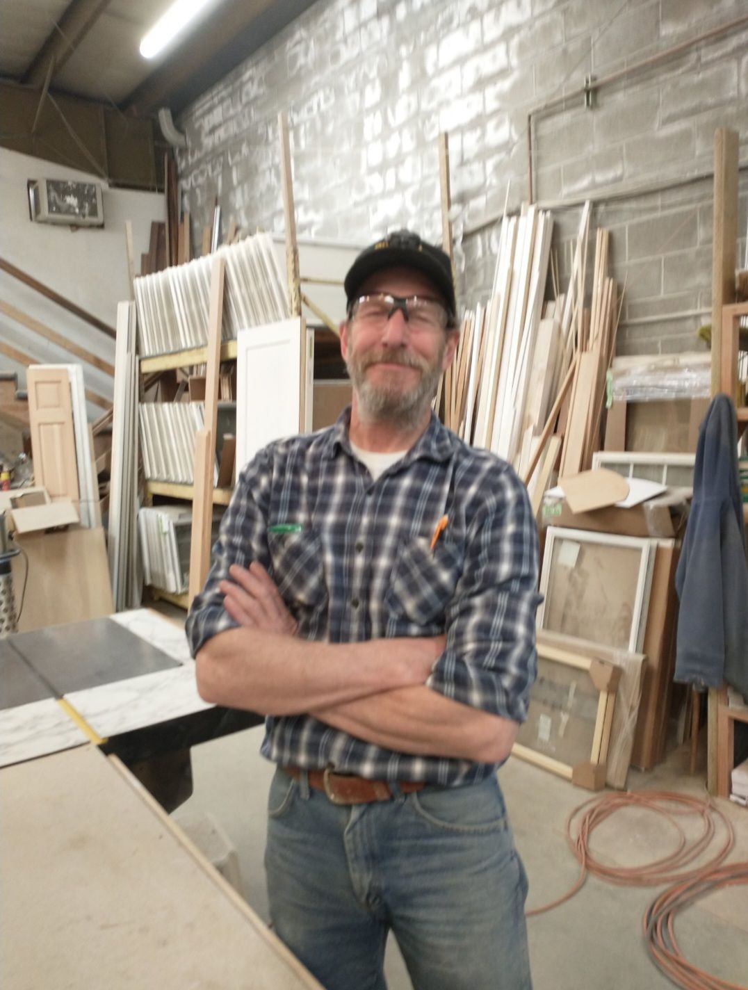A person in a plaid shirt, cap, and safety glasses stands with arms crossed in a woodworking shop filled with wood supplies.