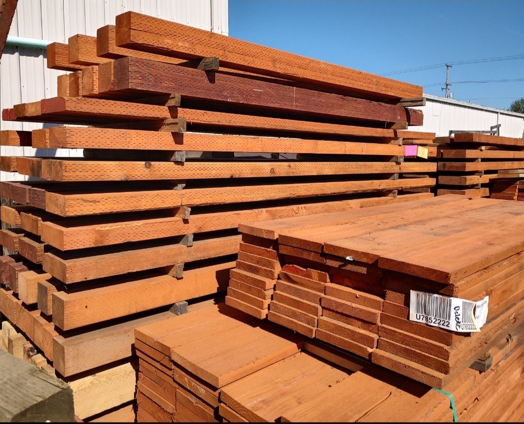 Stacks of brown, pressure-treated lumber beams and boards stored outdoors in a sunny supply yard